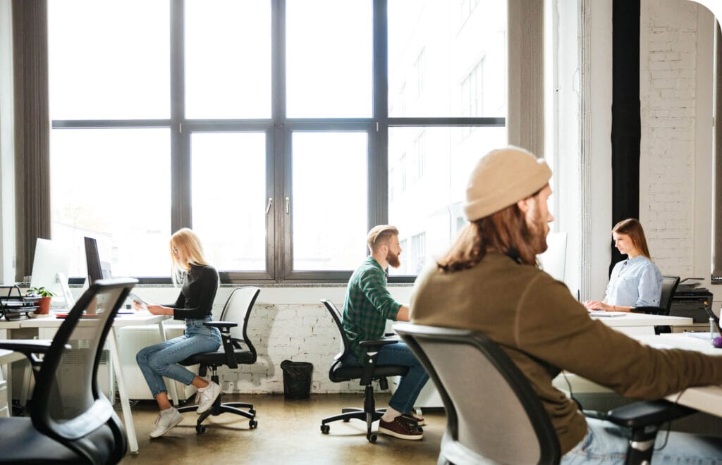 A bright, modern office with four people working on computers at desks, surrounded by large windows and a minimalist decor.