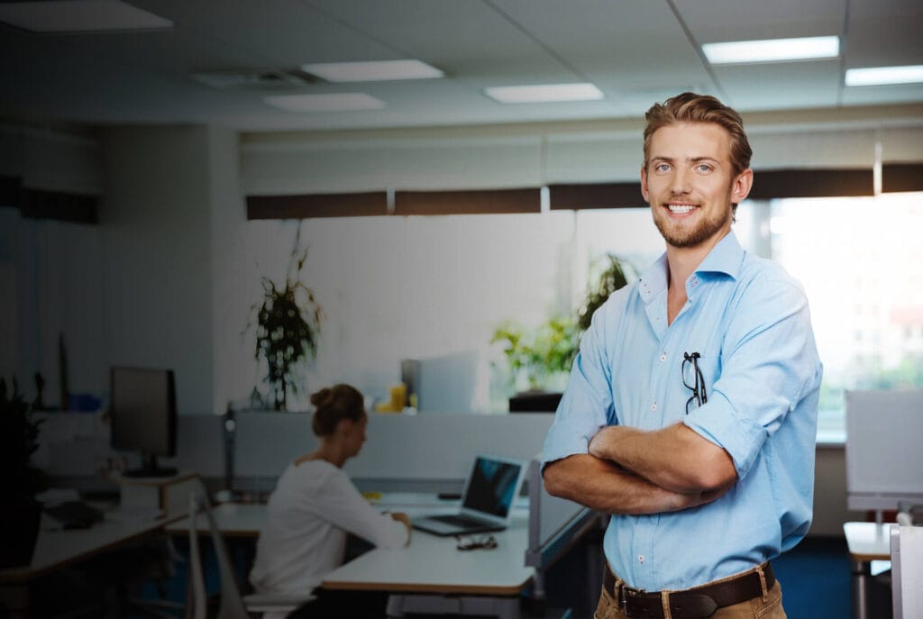 A confident man in a blue shirt stands with arms crossed in a bright, modern office with plants and a woman working at a desk.